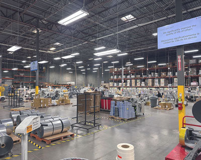 Large industrial warehouse with machinery, metal rolls, and stacked shelves under bright overhead lighting.